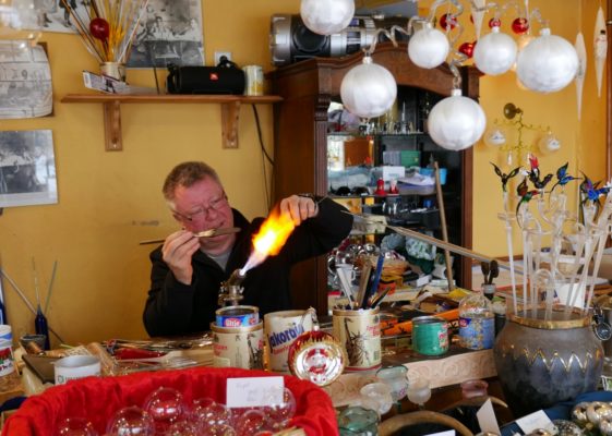 Ein Glasbläser sitzt hinter einer Gaslampe und erhitzt ein Glasobjekt. Auf seinem tisch die Arbeitsutensilien, über ihm weiße Glaskugeln für den Christbaum. Außerdem ein Gefäß mit mundgeblasenen Vögeln.