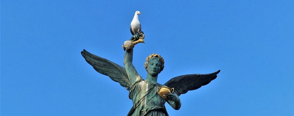 Eine weiß-graue Mittelmeermöwe steht hochoben auf einer Statue mit Flügeln. Der Himmel leuchtet intensiv blau.